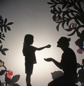 forest garden shadow theatre scene child feeds apple to mother by Jennie Pedley
