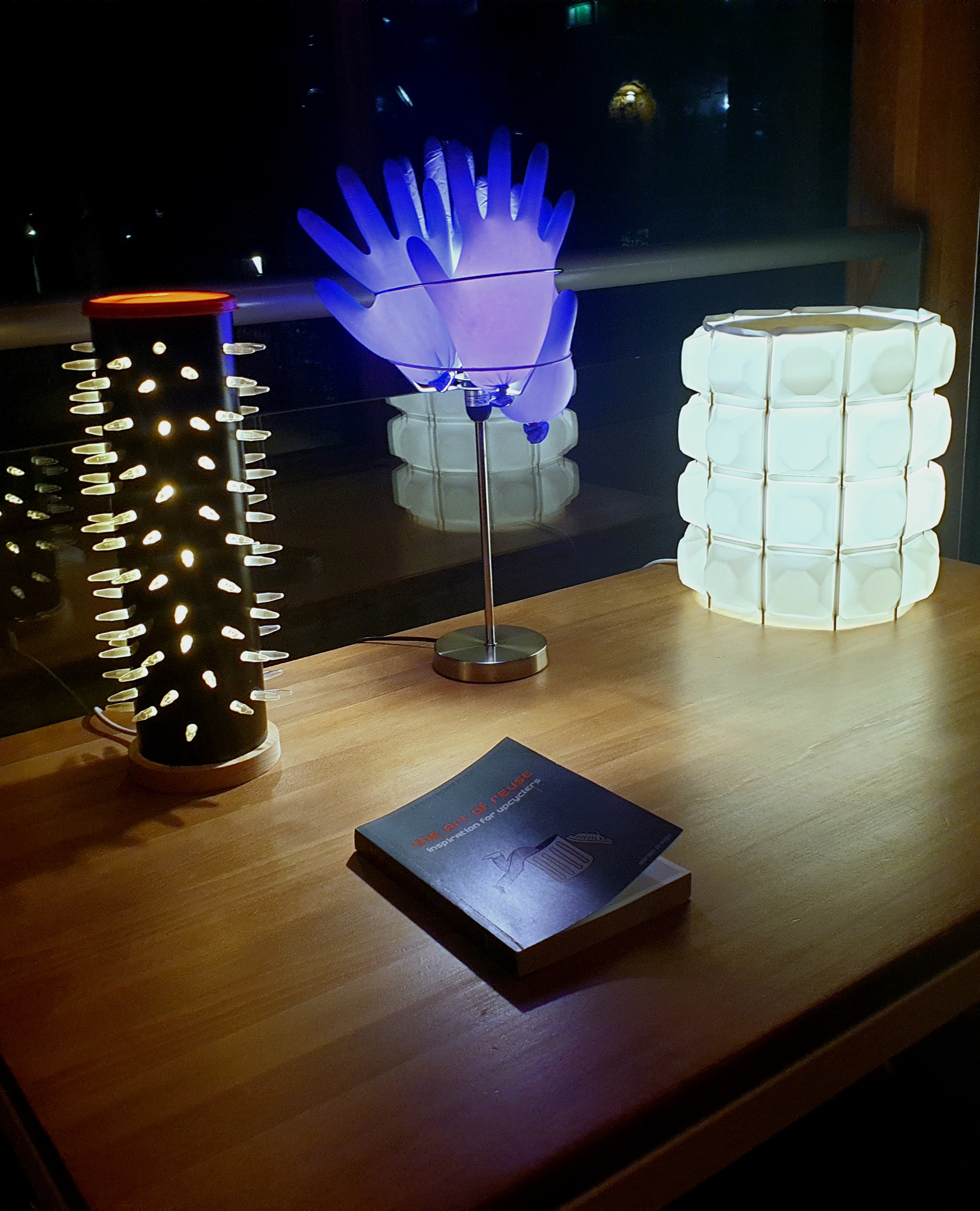 Inflated purple rubber gloves glow, on a stand, alongside a black tube with glowing spikes and a cylinder of white plastic squares all glowing on a table with a book in front called the Art of Reuse by Adrian Draigo